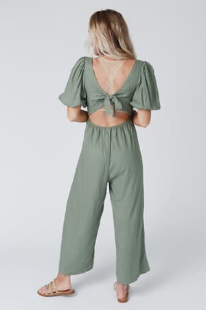 A woman stands facing away, wearing the Weekender Puff Sleeve Jumpsuit in olive with wide legs, an open back tied with a bow, and lace detailing. She has long, wavy blonde hair and is wearing sandals.