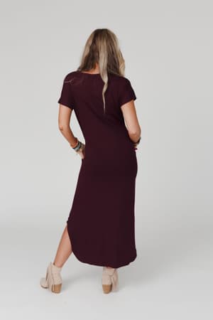 A woman with long, wavy hair stands with her back to the camera against a light background, wearing the Call It Comfort Tee Dress in plum—a relaxed fit, ankle-length style—paired with beige ankle boots.