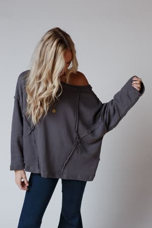 A woman with long, wavy blonde hair wears the Bailey Oversized Raw Edge Top in charcoal and dark jeans. She looks down, her arm extended to the side, standing against a plain light background.