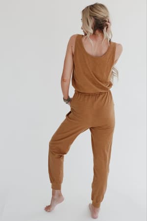 A woman with long, wavy blonde hair stands barefoot, facing away, modeling the Set to Go Textured Jumpsuit And Cardigan Set in camel against a plain white background.