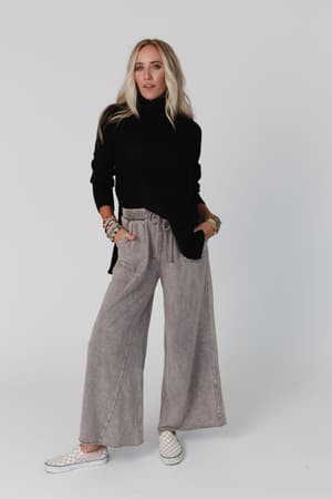 A woman stands against a plain background wearing the Danny Sweater in black, loose gray wide-leg pants, checkered slip-on shoes, and colorful bracelets, with her hands in her pockets.