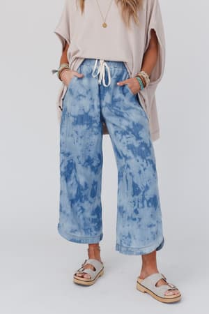 Wearing The Nest So Comfy Wide Leg Denim Pant in bleach dye with a beige loose top, platform sandals, and layered bracelets, a person stands hands-in-pockets against a plain background.