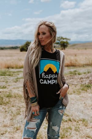 A woman with long blonde hair stands in a grassy field beneath a cloudy sky and mountains, wearing ripped jeans, jewelry, a black Happy Camper graphic tee, and the Dreamy Macramé Kimono - Mocha with tassel fringe.