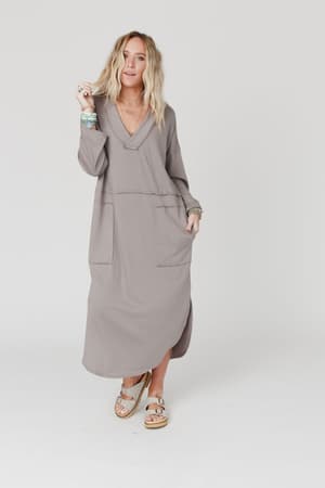 Woman with wavy blonde hair wears The Nest Meadow Long Sleeve Slit Maxi Dress - Coco with pockets, paired with sandals and bracelets, standing against a plain white background and smiling softly.