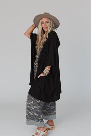 A woman with long blonde hair wears a wide-brimmed hat, black kimono-style top, camouflage pants, and sandals. She accessorizes with colorful bracelets and drapes The Nest Boho Essential Hooded Ribbed Sweater - Black over her shoulders.