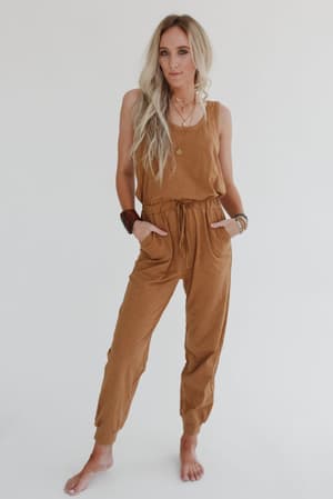 A woman with long blonde hair stands barefoot against a white background, wearing the Set to Go Textured Jumpsuit and Cardigan Set in Camel. She completes the look with layered necklaces and bracelets, her hands tucked in her pockets.