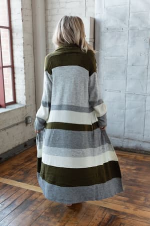 A woman with long blonde hair stands indoors on a wooden floor, facing away, wearing The Nest So Cozy Oversized Striped Knit Duster in Olive Multi. The cozy room has white brick walls and large windows.