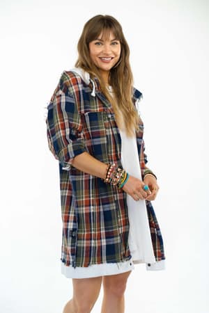 A woman with long brown hair smiles as she wears the Flannel Layers Hooded Jacket in Navy Multi over a white dress, accessorized with colorful bracelets, standing against a plain white background.