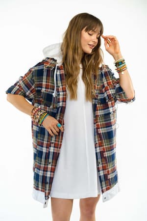 A woman poses confidently against a white background, wearing the Flannel Layers Hooded Jacket - Navy Multi over a white dress. She has long, wavy hair and colorful bracelets on both wrists.
