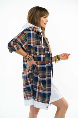 A woman in profile wears the Flannel Layers Hooded Jacket - Navy Multi over a white hoodie and dress. She has long hair with bangs and colorful bracelets on both wrists.