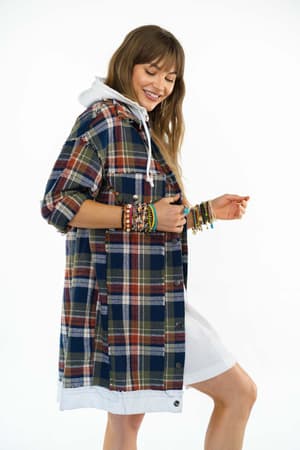 A smiling woman stands against a plain white background, wearing the Flannel Layers Hooded Jacket in Navy Multi over a white hooded dress and accessorized with multiple colorful bracelets.