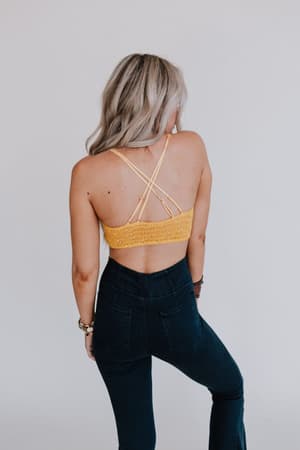 A person with long, wavy blonde hair faces away from the camera, wearing the Dainty Daisy Crochet Bralette in yellow and high-waisted dark jeans against a plain light background.