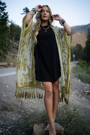 A woman stands barefoot on a tree stump by the roadside in a black dress and the Crushin' On You Velvet Kimono - Olive, featuring tassel details and bracelets. Trees and mountains are seen in the background.