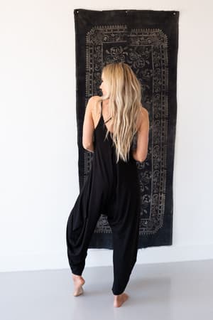 A woman with long blonde hair stands barefoot with her back to the camera, wearing The Perfect Harem Jumpsuit - Black in front of a decorative black tapestry on a white wall.