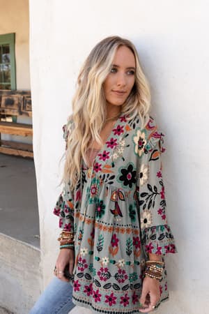 A woman with long, wavy blonde hair stands against a white wall, wearing the Leona Floral Print Tunic in Sage and several bracelets, looking thoughtfully to the side.