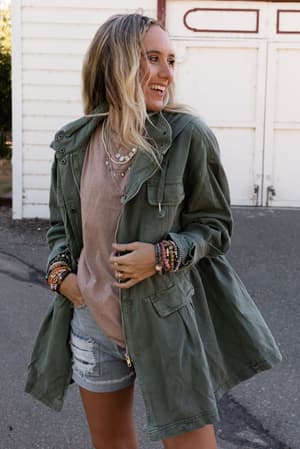 A smiling woman with long blonde hair wears the Layers On Layers Perfect Boho Parka in olive over a beige t-shirt and ripped denim shorts, accessorized with layered necklaces and bracelets, standing outside a white building.