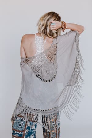 A woman faces away from the camera, wearing the Dreamy Macramé Kimono in mocha over a white lace halter top and patterned pants. Her blonde hair is styled in loose waves.