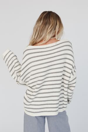 A woman with wavy blonde hair, seen from behind, wears the All My Stripes Oversized Sweater in ivory gray paired with light gray pants.