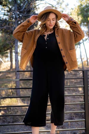 A woman in a tan jacket and hat stands outdoors by a metal railing, looking down and touching her hat brim, showing off the relaxed, oversized silhouette of the Simply Comfort Jumpsuit - Black. Trees are visible in the background.