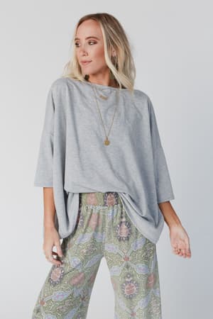 A woman wears The Nest Cozy Cool Oversized Tee - Heather Gray with patterned wide-leg pants, standing against a plain light background. She looks to the side, hand on hip, with wavy blonde hair and a long necklace for a relaxed boho vibe.