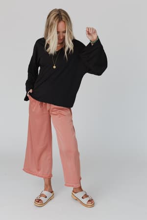 A blonde woman wears The Nest Love It V Neckline Sweater Top in black, peach wide-leg pants, white sandals, and a necklace. She stands against a plain background, looking down with one hand in her pocket and the other raised.