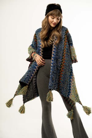 A woman wearing a black headband, cobalt Desert Dreams Crochet Ruana with tassels, black top, and gray flared pants stands against a white background, looking down with a gentle smile—a chic addition to any boho closet.