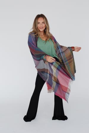 A woman with long blonde hair wears a green top, black pants, colorful bracelets, and the Cozy Plaid Pocket Ruana Wrap - Fuchsia. She stands against a plain white background, smiles slightly, and gestures with one hand.
