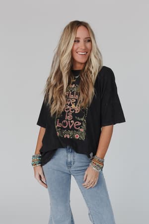 A woman with long, wavy blonde hair wears the Share The Love Graphic Tee in charcoal, paired with light blue jeans and colorful beaded bracelets, standing and smiling against a plain background—ideal for a boho-inspired closet.