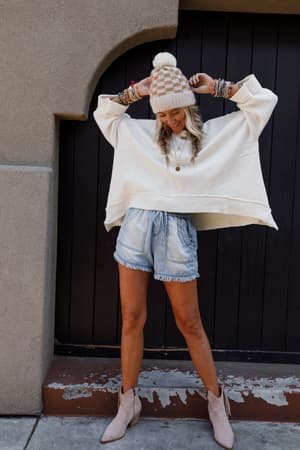 A woman stands in front of a dark wooden door, smiling with arms raised, wearing the Dana Oversized Diamond Texture Sweater in Ivory, a beige knit hat, light denim shorts, ankle boots, and multiple bracelets.