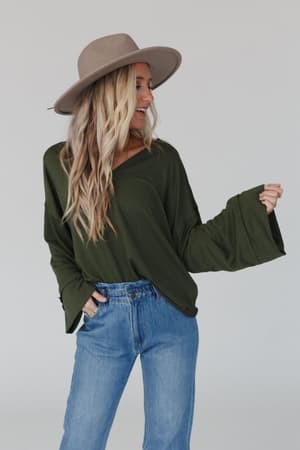 A woman with long blonde hair smiles as she looks to the side, wearing The Nest Love It V Neckline Sweater Top in olive, styled with a wide-brimmed beige hat and blue jeans, and keeping one hand in her pocket.