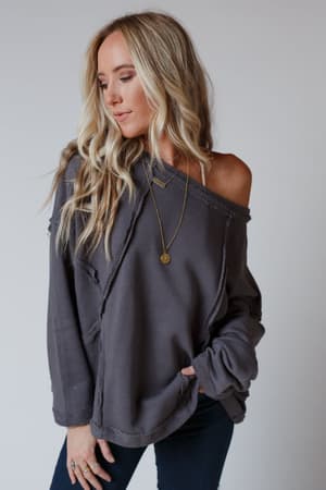 A woman with long, wavy blonde hair wears the Bailey Oversized Raw Edge Top in charcoal with dark jeans, standing against a plain light background and looking down with one hand in her pocket for a relaxed boho style.