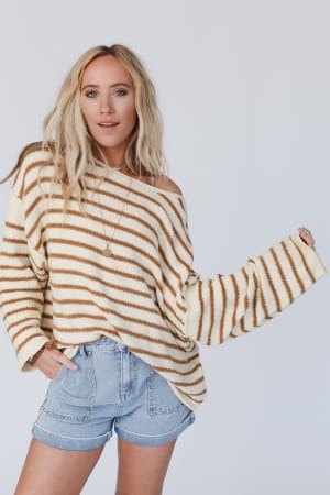 A woman with long blonde hair models the All My Stripes Oversized Sweater in mustard, featuring loose sleeves, paired with light blue denim shorts. She poses against a plain light background, gazing at the camera with a relaxed expression.