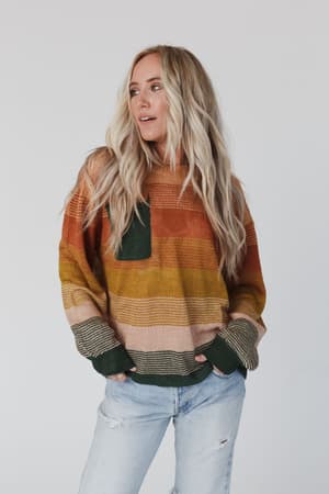 A woman with long, wavy blonde hair wears the Going Places Gradient Stripe Sweater - Mustard and light blue ripped jeans, standing against a plain white background and looking slightly to her left.