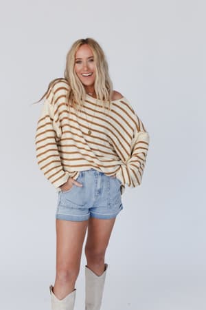 A woman with long blonde hair wears the All My Stripes Oversized Sweater in mustard, paired with denim shorts and white knee-high boots. She stands against a plain light background, smiling with her hands in her pockets.