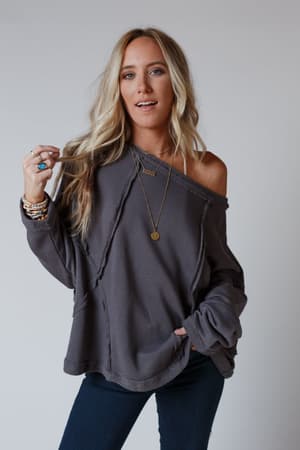 A woman with long blonde hair wears the Bailey Oversized Raw Edge Top in charcoal with dark jeans, layered necklaces, bracelets, and rings, posing confidently against a plain light background.