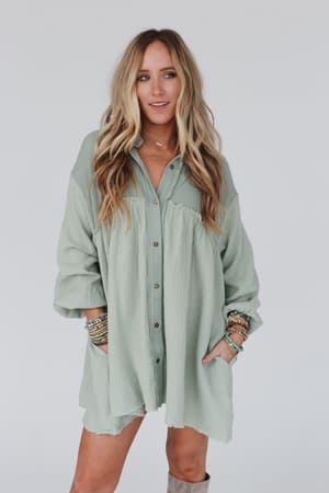 A woman with long, wavy blonde hair wears the Sunday Kind Of Love Dress in sage, an oversized boho style button-up with rolled sleeves, accessorized with stacked bracelets, standing against a plain light background.