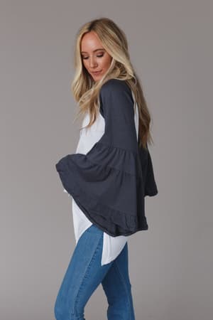 A woman with long blonde hair models the Bell Sleeve Baseball Tee - Charcoal with dark gray boho-chic bell sleeves, paired with blue jeans, standing in profile against a plain gray background.