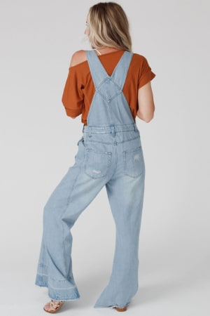 A person with long blonde hair stands barefoot and faces away from the camera, wearing a rust short-sleeve top and the Milly Distressed Hem Wide Leg Overall - Denim, creating a subtle boho vibe against a plain background.