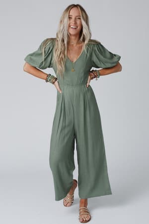 A smiling woman with long blonde hair wears the Weekender Puff Sleeve Jumpsuit - Olive, styled with layered necklaces, bracelets, and sandals, standing against a plain light background.
