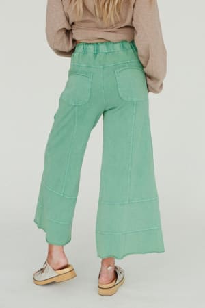 A person stands facing away in boho style, wearing the Lucky One Wide Leg Pant - Green with back pockets and a beige long-sleeve top. Their long blonde hair and beige slip-on platform shoes complete the look.