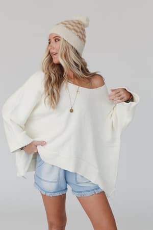 A woman stands against a plain light background, looking to the side, wearing the Dana Oversized Diamond Texture Sweater in Ivory, paired with light denim shorts and a white beanie with a tan pattern.