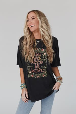 A woman with long, wavy blonde hair smiles in the Share The Love Graphic Tee - Charcoal featuring floral graphics and "All We Need Is Love," styled with beaded bracelets and light blue jeans for a perfect boho-inspired look.