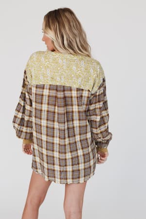 A woman with long, wavy blonde hair faces away, modeling the Better Together Mixed Print Top - Mustard, which features a yellow floral yoke and brown plaid body, set against a plain white background.