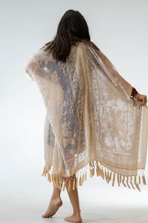 A person with long dark hair stands barefoot, facing away, wearing the Crushin' On You Velvet Kimono in nude. The lightweight fabric drapes loosely and features fringe and tassel details for a delicate boho look.