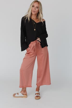 A woman with long blonde hair and a relaxed pose stands against a plain background, wearing The Nest Love It V Neckline Sweater Top in black, peach wide-leg pants, white sandals, and layered gold necklaces.