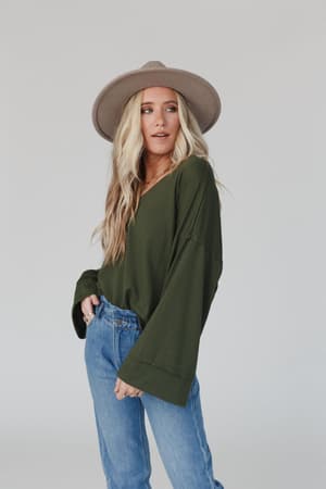 A woman with long blonde hair in a wide-brimmed beige hat and The Nest Love It V Neckline Sweater Top in olive, paired with blue jeans, stands against a light gray background, looking slightly to the side.
