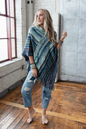 A woman in ripped jeans and a vibrant Madeleine Poncho - Blue with fringe stands indoors by large windows on a wooden floor, her wrist stacked with bracelets and her long blonde hair in loose waves.