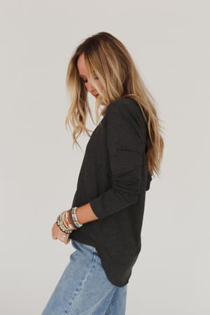 A woman with long blonde hair, wearing the Ryder Long Sleeve Bralette Tee in Charcoal and light blue jeans with colorful beaded bracelets, stands in profile against a plain white background.