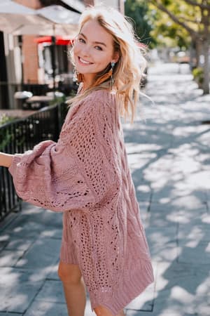 A smiling woman walks outdoors on a sunny sidewalk, turning back to the camera, wearing the Golden Hour Bubble Sleeve Sweater in mauve. Trees and buildings line the street in the background.