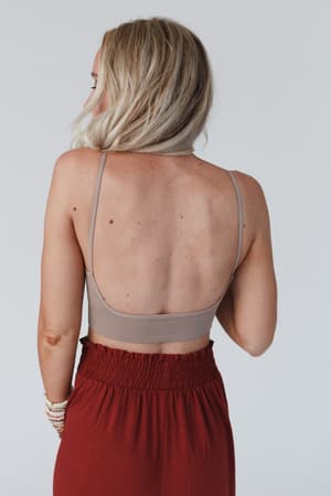 A woman with blonde hair faces away from the camera, wearing the Dora Low Back Seamless Bralette in Mocha and high-waisted, rust-colored pants against a plain light background.